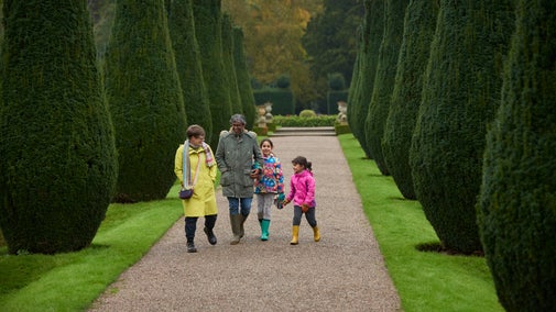 A family consisting of a man, a woman, and two young girls walking down a path in Erddig's garden during autumn. They are smiling and laughing, surrounded by bushes.